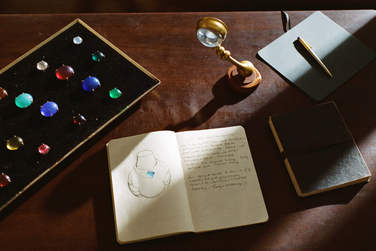 A gemstone consultant's desk: a velvet tray of varied polished gems, a small sphere on a stand, a hand-drawn ring sketch, an open notebook, brass loupe and pen on dark walnut